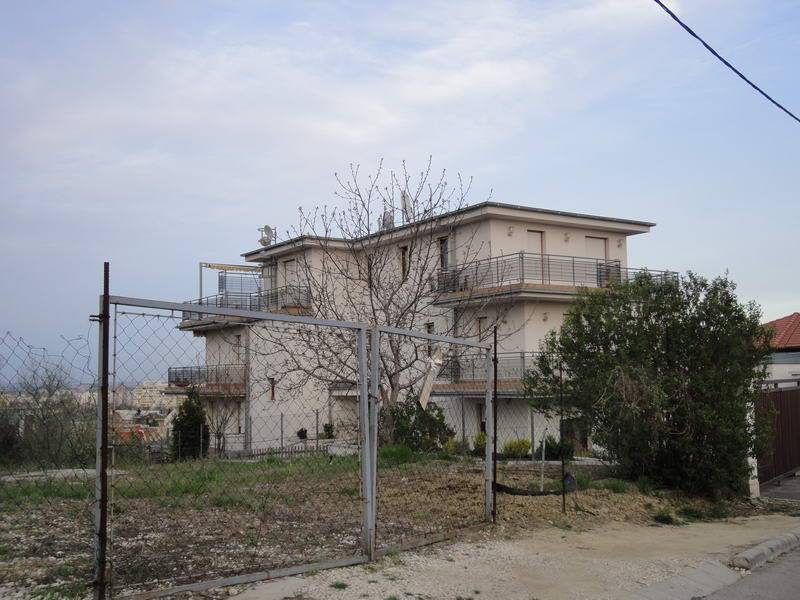 A pale multi-storey building with broad balconies is viewed from beyond a chain-link fence. A bare tree and leafy shrubs partly obscure the facade. An open patch of rough ground lies in the foreground, and a dark cable crosses the sky near the upper right.