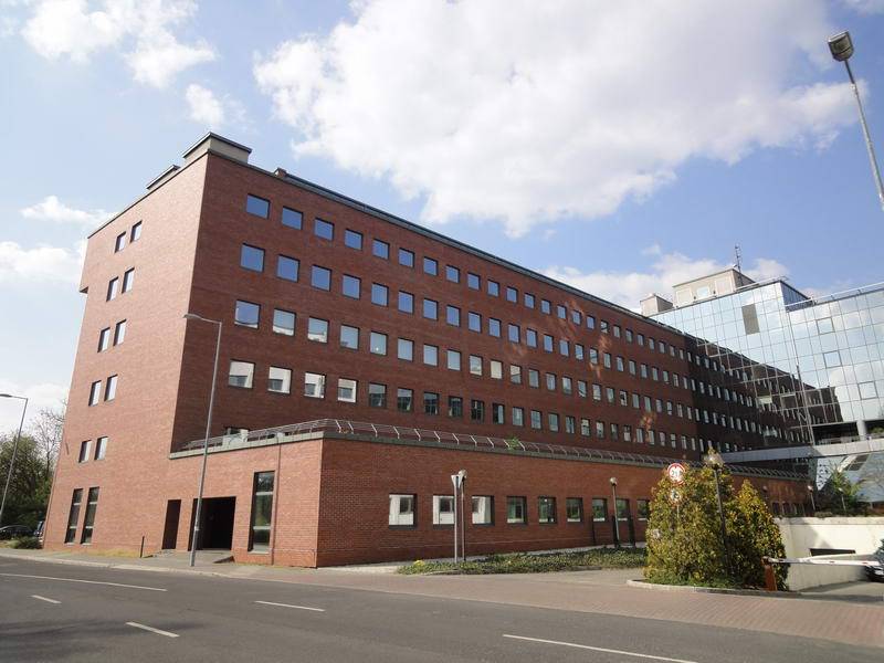 A low-angle street view shows a long multi-storey building with a red brick facade and rows of rectangular windows. A reflective glass wing adjoins the brick section on the right. A paved road and sidewalk cross the foreground beneath a partly cloudy sky.