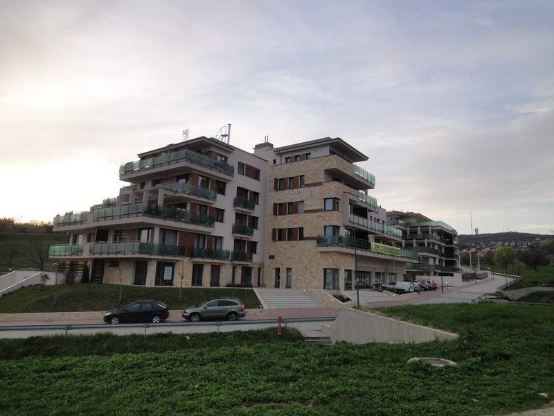 The image shows a multi-storey building with staggered balconies and glass railings. Pale masonry and brick sections form the facade, with cars parked beside a road along the front. A grassy slope fills the foreground under an overcast sky.