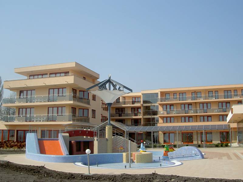 The image shows a large light-colored multi-storey complex with repeated balconies and rows of windows. A paved courtyard in front contains a curved blue-and-orange structure and several low circular features. A pale canopy-like structure rises between the building sections under a clear sky.