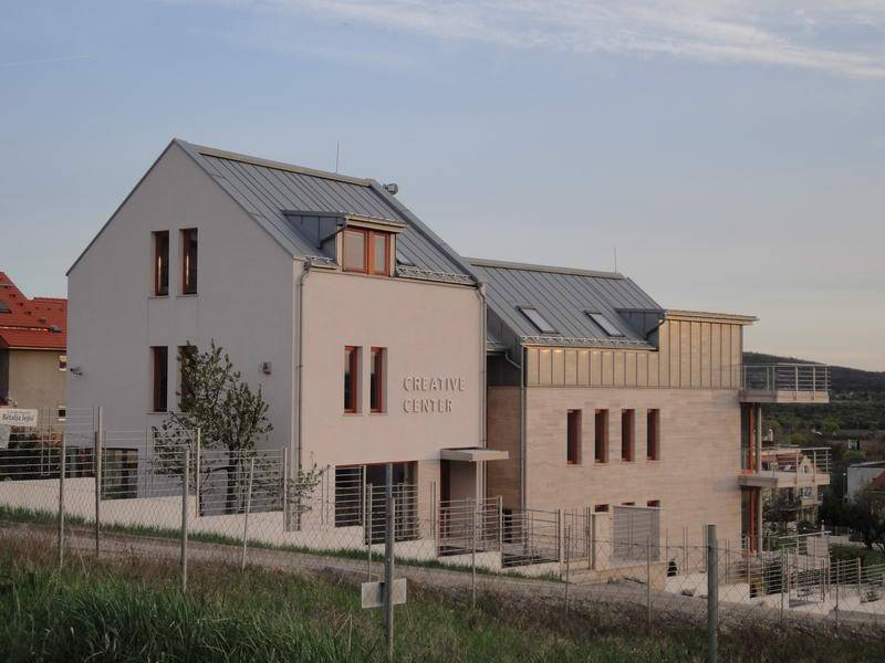 A pale multi-level building with several roof sections is viewed from across a fenced slope. The words “CREATIVE CENTER” are visible on the front wall. Narrow windows, a metal fence, and a terrace or balcony structure appear along the lower and right sides.