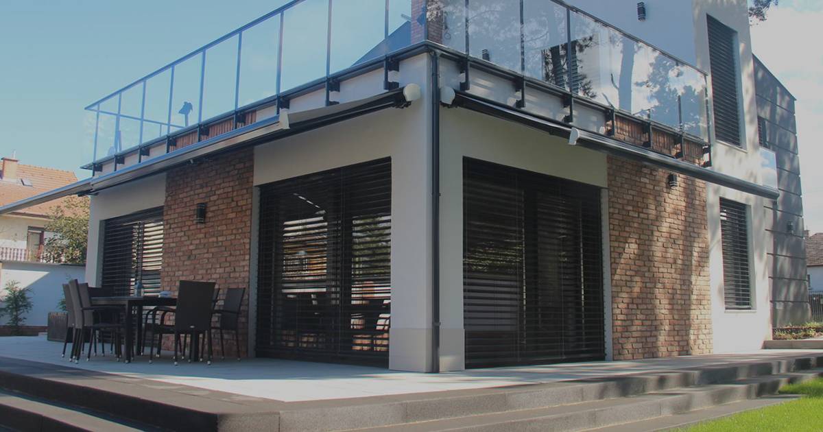 An angled exterior view shows a two-storey building with brick walls, light-colored wall sections, and large dark horizontal louvered coverings over the lower windows. A glass railing runs along the upper level, and a patio table with chairs sits to the left. Wide concrete steps and a patch of lawn occupy the foreground.