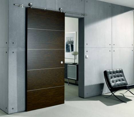 A dark wood sliding door is partly open on an exposed metal track mounted above the opening. Thin horizontal light-colored lines cross the door surface, and a small round fitting is visible near its lower right area. The surrounding room has gray concrete walls, a black lounge chair, and a glimpse of a furnished room through the opening.