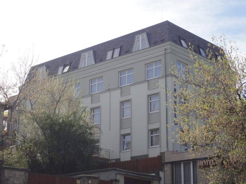 A pale multi-storey facade with rows of windows sits beneath a broad dark mansard-style roof. Bare and budding trees partly obscure the building. The word “HOTEL” is visible on a sign at the lower right, with a fence and stone wall along the bottom edge.