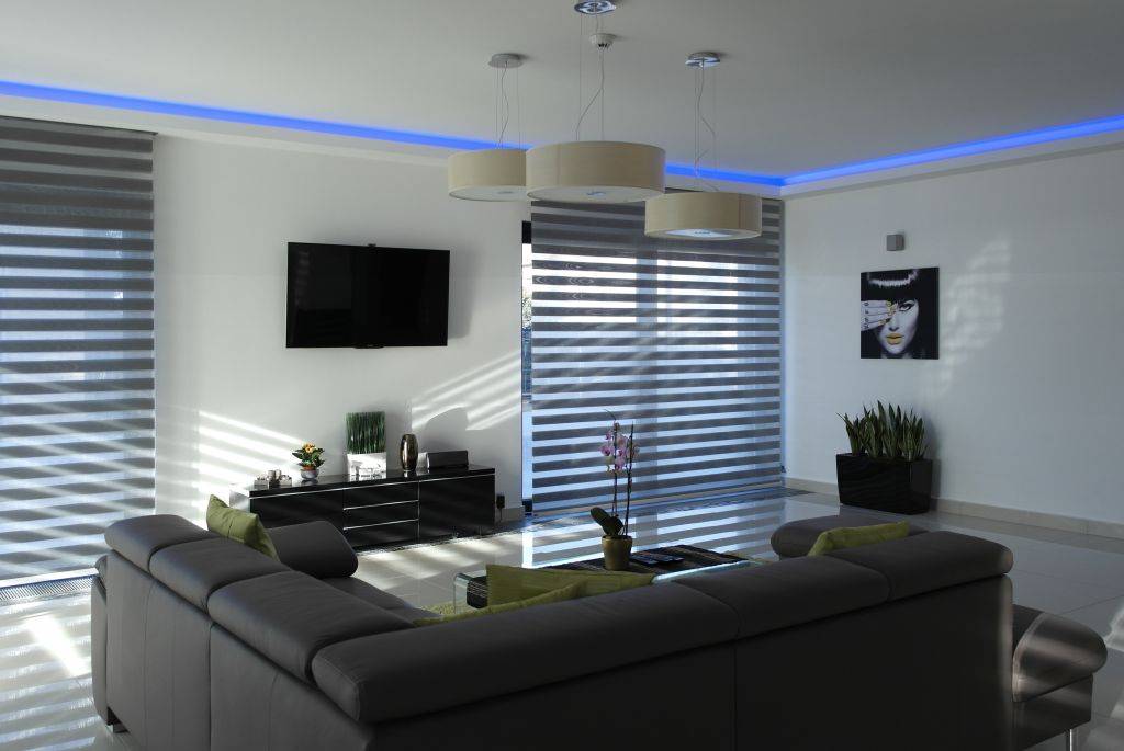 A bright living room is shown with two large windows fitted with alternating horizontal gray and white bands. A gray sectional sofa fills the foreground, facing a wall-mounted television and low black cabinet. Blue accent lighting follows the ceiling perimeter, and plants, hanging lights, and wall art are visible.