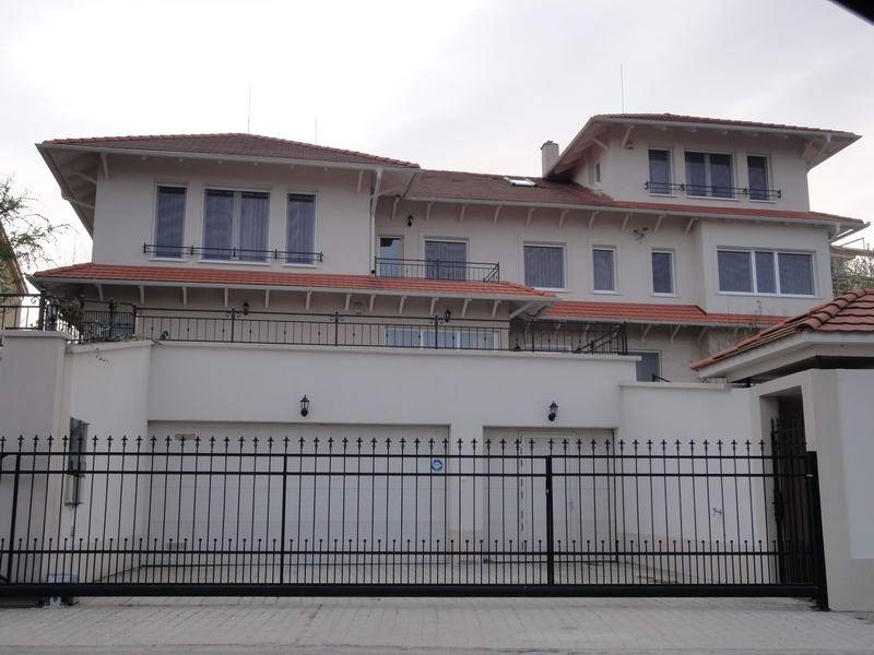 A long, white multi-level building with several connected roof sections is viewed from outside a black metal gate. Red roof tiles cover the main sections, and railings run along the visible upper balconies. A pale paved drive fills the foreground, with a small portion of a neighboring roof at the right.